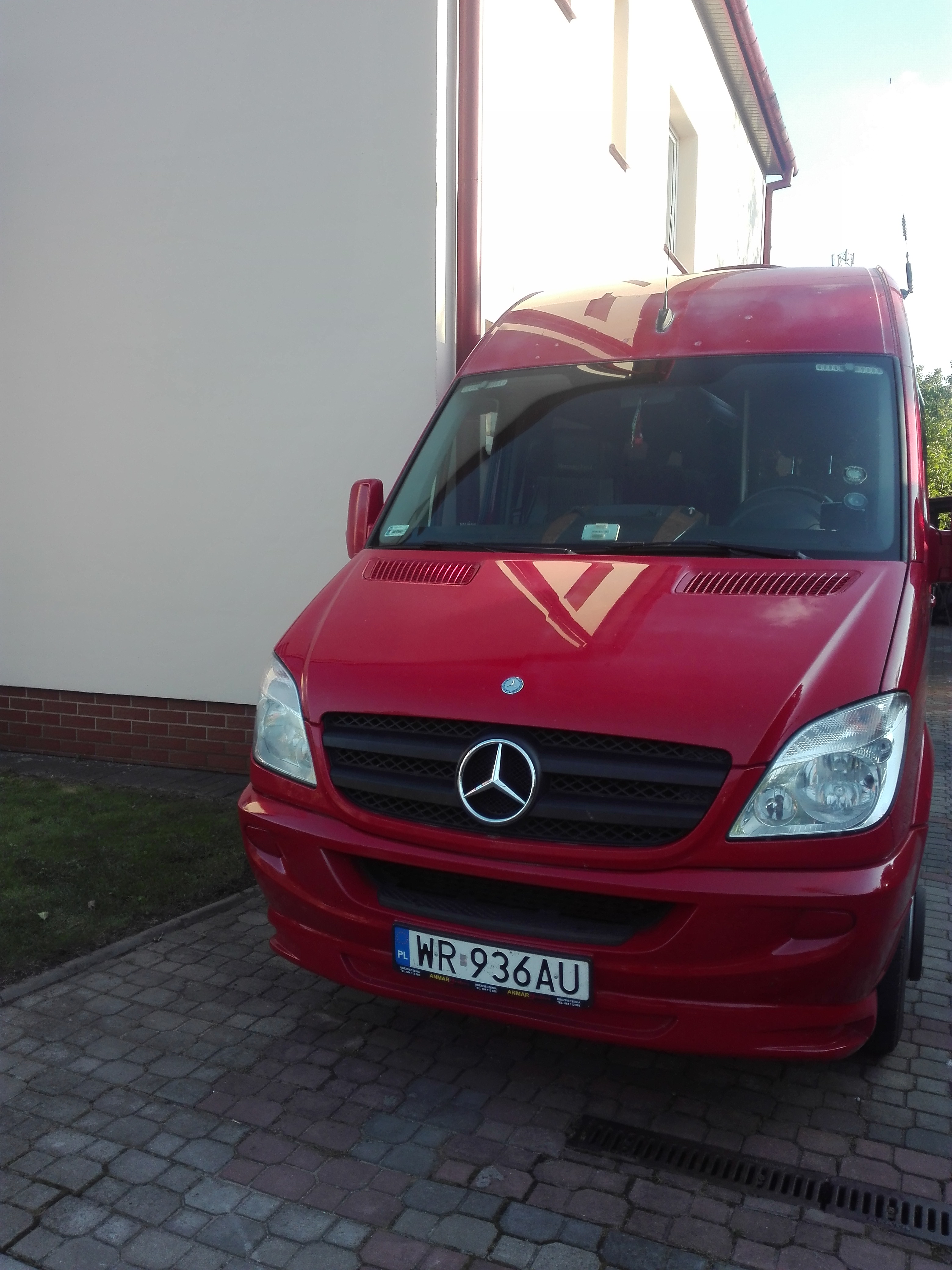 Czerwony bus Mercedes-Benz Sprinter zaparkowany na brukowanej nawierzchni przed budynkiem z białą elewacją i czerwonym dachem.