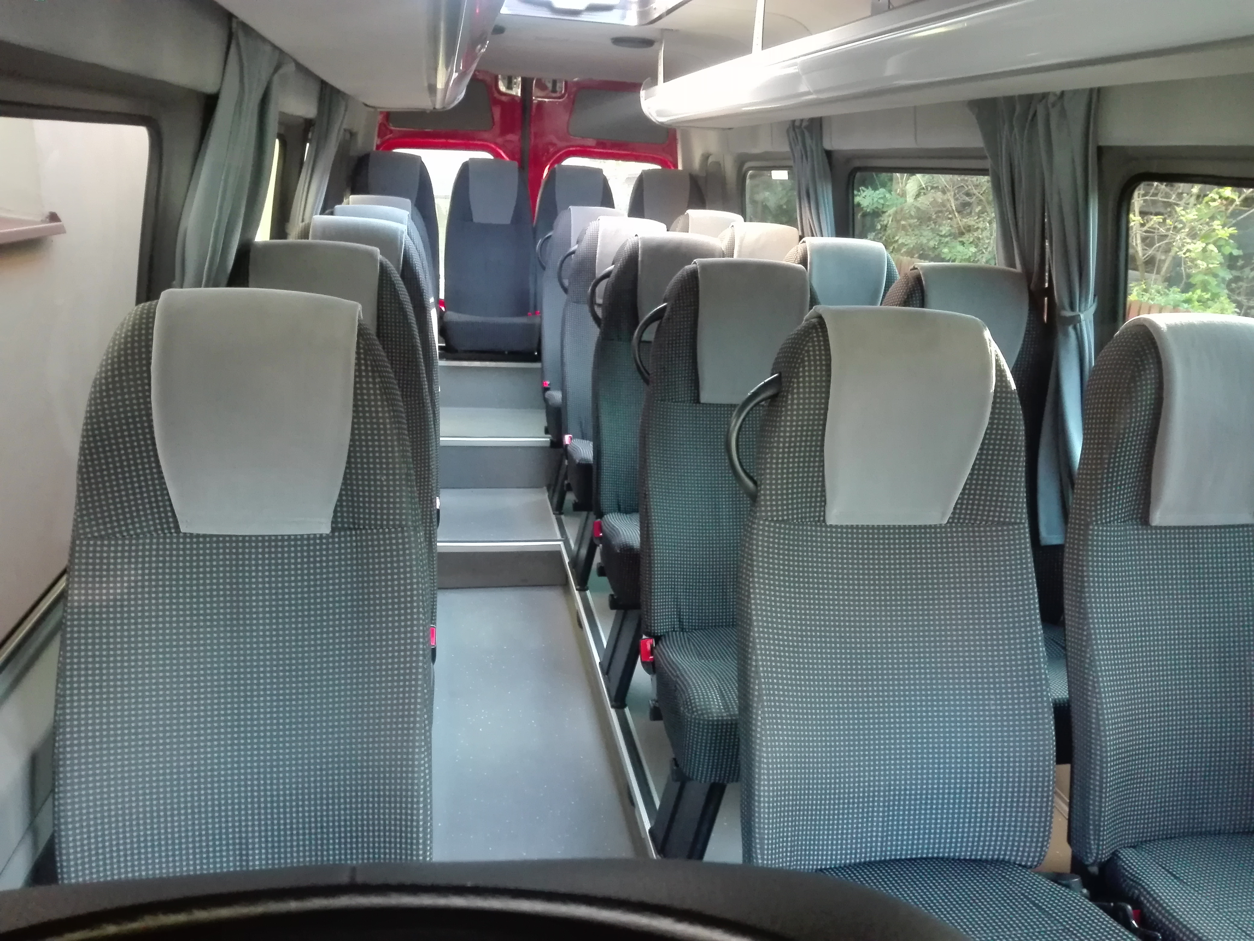 Wnętrze autobusu z rzędami szarych foteli z zagłówkami, widok z perspektywy kierowcy, zasłonięte okna z boku.