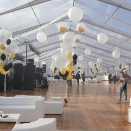 Wnętrze dużego, przezroczystego namiotu eventowego z drewnianą podłogą, białymi sofami i dekoracjami z białych, czarnych i złotych balonów, nagłośnienie w tle.