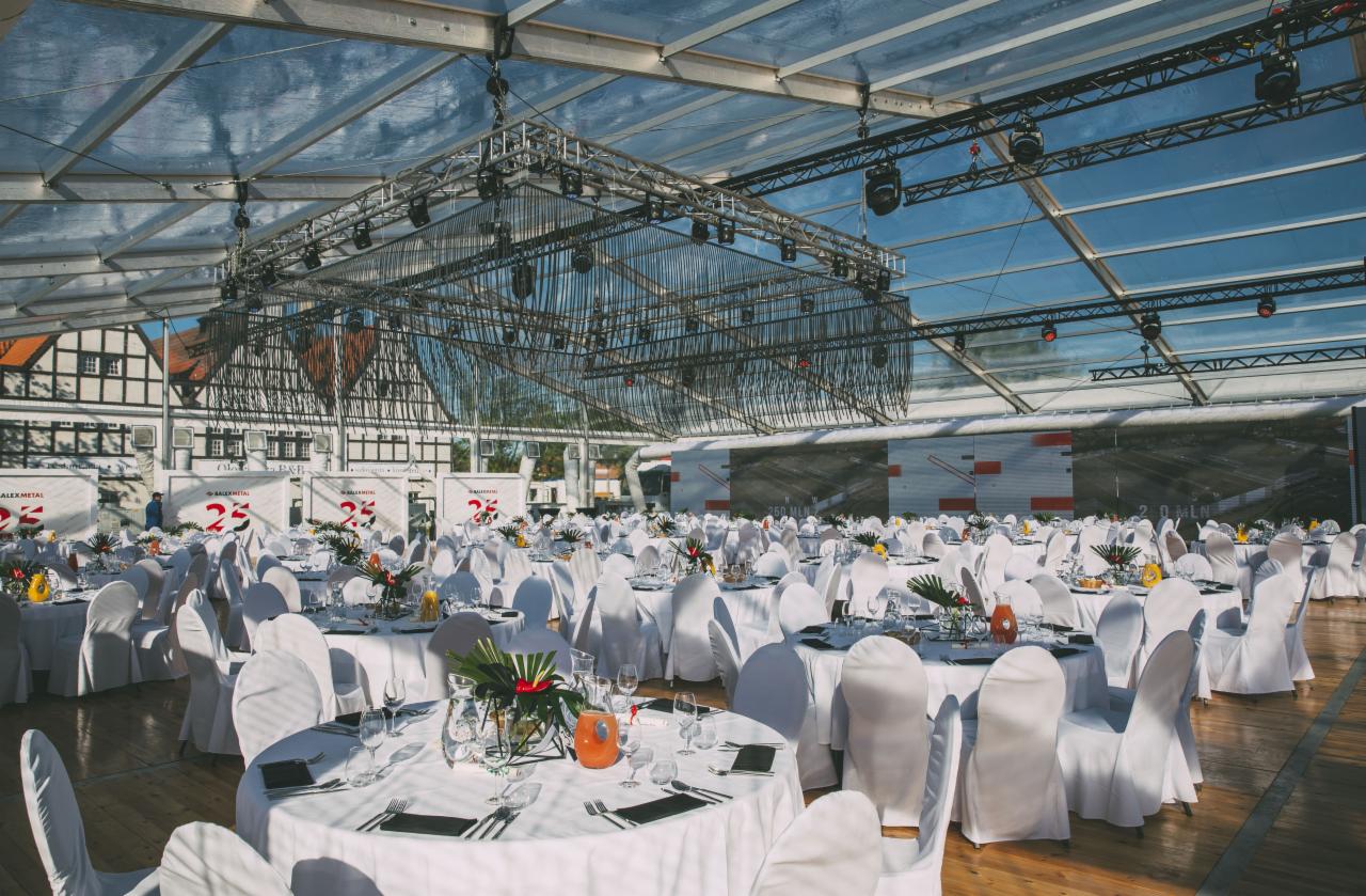 Uroczysta sala bankietowa w przeszklonym namiocie z dekoracjami kwiatowymi na stołach, oświetleniem technicznym pod sufitem i parkietem na podłodze.