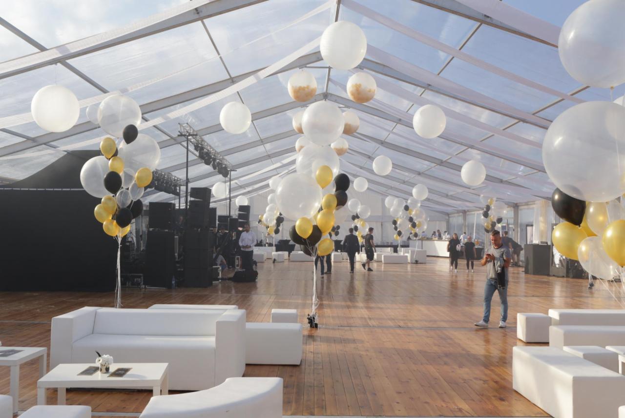 Wnętrze dużego, przezroczystego namiotu eventowego z drewnianą podłogą, białymi sofami i dekoracjami z białych, czarnych i złotych balonów, nagłośnienie w tle.