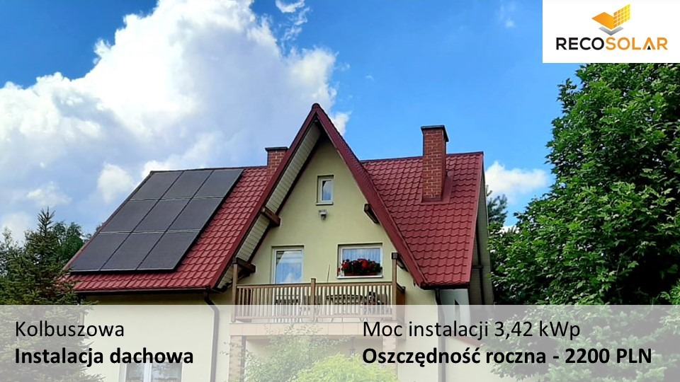 Dom jednorodzinny z czerwoną dachówką, na której zamontowano panele słoneczne. Widoczne błękitne niebo z białymi chmurami i logo firmy RecoSolar w prawym górnym rogu.