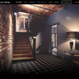 Wnętrze Aparthotelu Sorau w Żarach: schody z drewnianymi stopniami, ceglana ściana, czarno-biała podłoga w szachownicę oraz widok na pokój z lampą i fotelem.