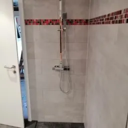 Nowoczesna kabina prysznicowa z deszczownicą i odpływem liniowym, wykończona szarymi płytkami z dekoracyjnym pasem mozaiki w odcieniach czerwieni i czerni.