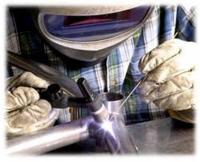 Pracownik w masce ochronnej spawa metalowy element metodą TIG, widoczny intensywny łuk elektryczny i iskry.