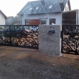 Czarna brama wjazdowa przesuwna z motywem drzewa, zamontowana między kamiennymi słupkami; w tle dom z szarą elewacją i zaparkowany samochód dostawczy.