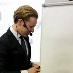 Prelegent w garniturze i mikrofonie nagłownym, pochylony, z whiteboardem w tle, prawdopodobnie podczas szkolenia lub prezentacji.