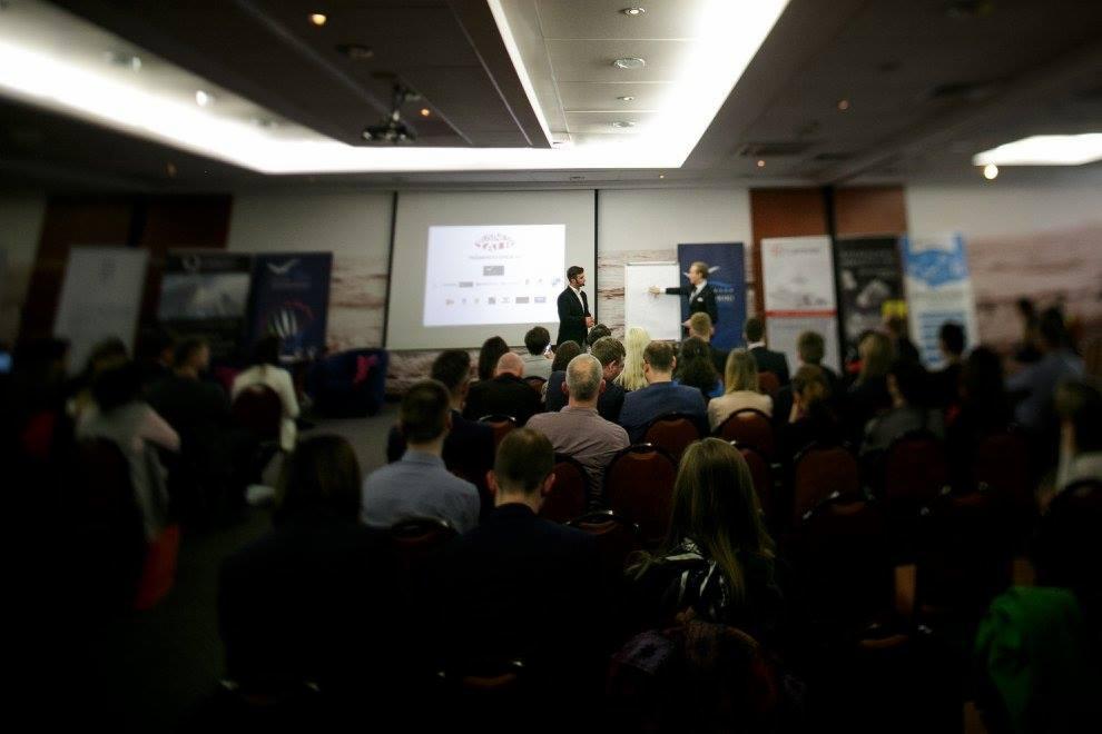 Prelegenci prowadzą prezentację dla publiczności siedzącej w rzędach krzeseł na sali konferencyjnej; projektor wyświetla slajdy na ekranie.