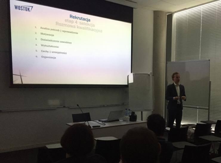 Prelegent w garniturze prowadzi prezentację o rekrutacji, wyświetlaną na ekranie projektora w sali konferencyjnej z widocznymi rzędami krzeseł i uczestnikami.