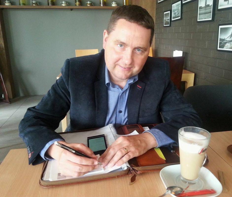 Mężczyzna w marynarce, siedzący przy stole, pisze w otwartym kalendarzu, obok szklanka latte i dekoracje ścienne.