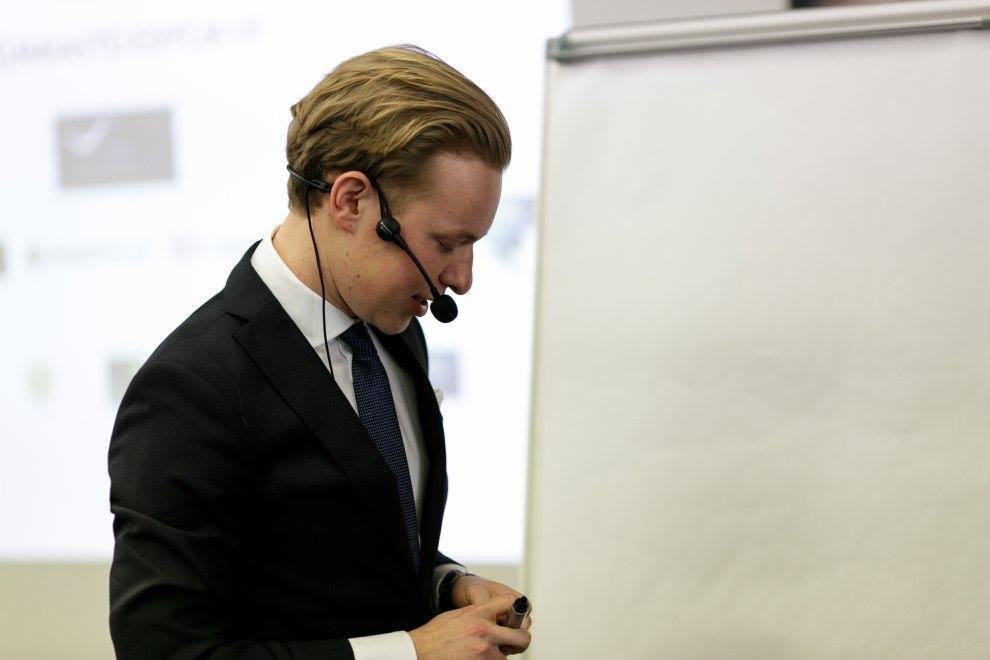 Prelegent w garniturze i mikrofonie nagłownym, pochylony, z whiteboardem w tle, prawdopodobnie podczas szkolenia lub prezentacji.