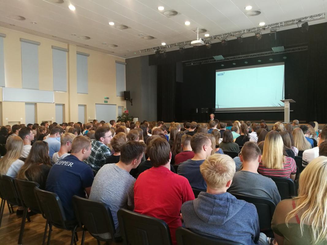 Sala konferencyjna wypełniona uczestnikami podczas wykładu, na scenie prelegent prezentuje treść na ekranie.