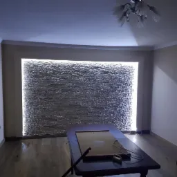 Ściana w salonie z dekoracyjną, podświetloną okładziną kamienną, widoczny stół i meble.