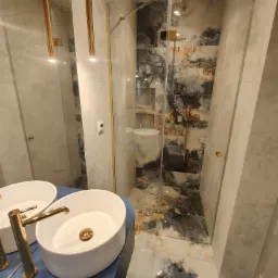 Nowoczesna łazienka z dwoma białymi umywalkami nablatowymi z bateriami w kolorze złotym, kabiną prysznicową ze szklanymi drzwiami i płytkami imitującymi marmur w odcieniach szarości, czerni i złota.