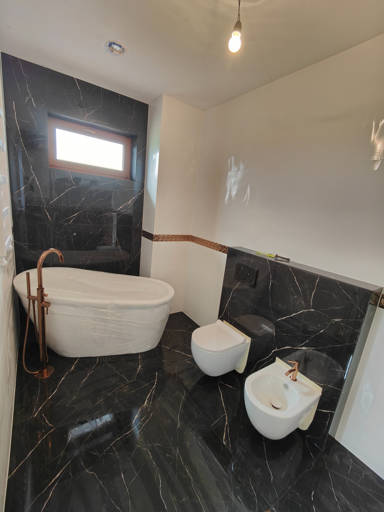 Nowoczesna łazienka z czarnymi marmurowymi płytkami, białą ceramiką sanitarną i armaturą w kolorze różowego złota, z wanną owiniętą folią ochronną.