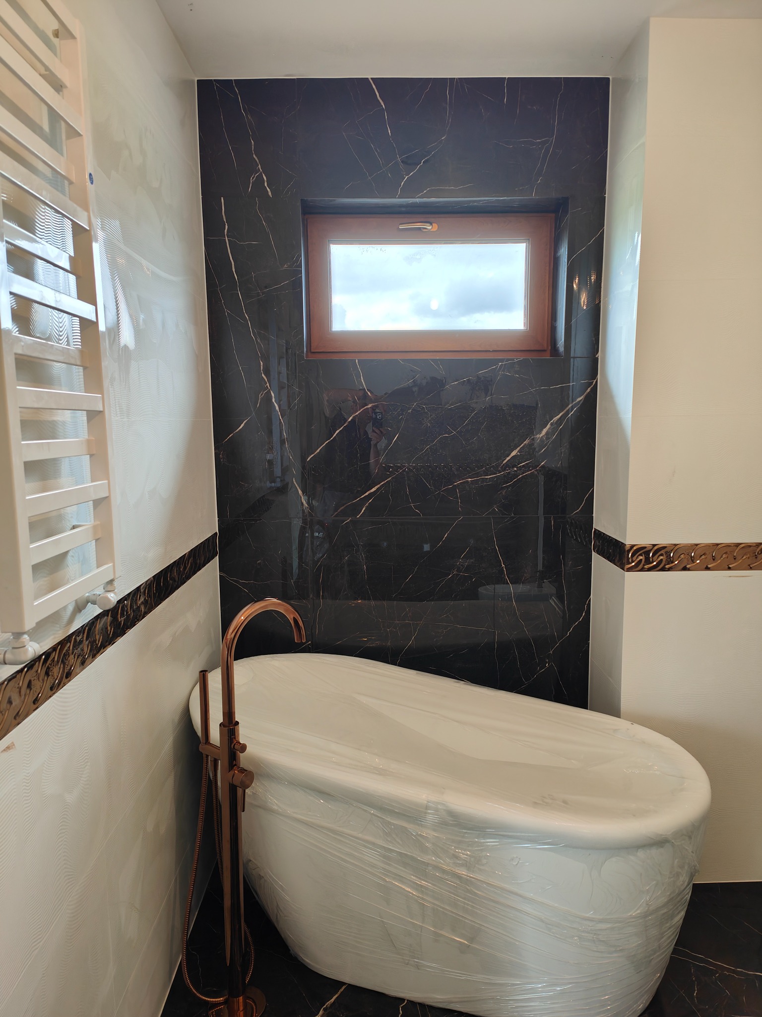 Nowoczesna łazienka z białą wanną owalną zabezpieczoną folią, baterią w kolorze różowego złota i ścianami wyłożonymi białymi i czarnymi płytkami imitującymi marmur, widoczne okno z drewnianą ramą.