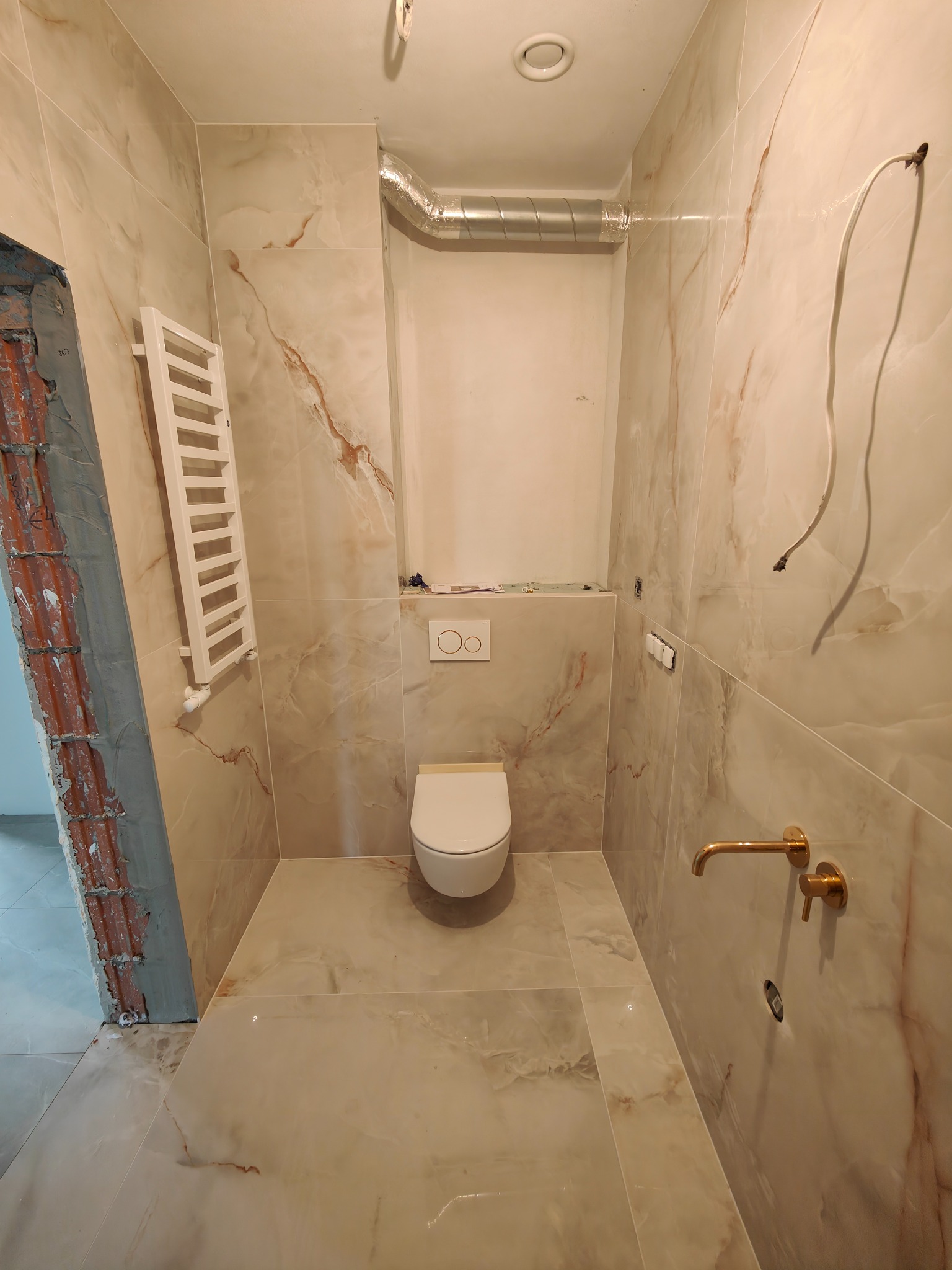 Wnętrze łazienki w trakcie remontu, wykończone płytkami imitującymi marmur, widoczna toaleta, grzejnik drabinkowy, złota bateria ścienna i niezamaskowany przewód elektryczny.