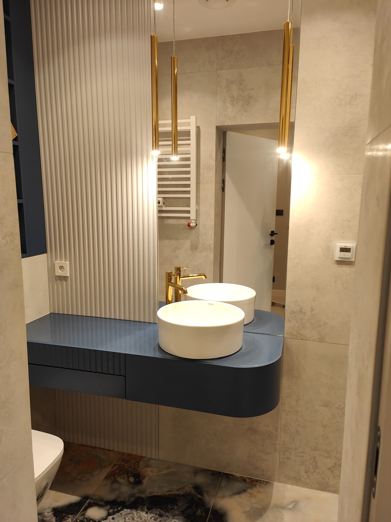 Nowoczesna łazienka z dwoma okrągłymi umywalkami na niebieskim blacie, złotą armaturą i wiszącymi lampami, widok na grzejnik drabinkowy i dekoracyjne panele ścienne.