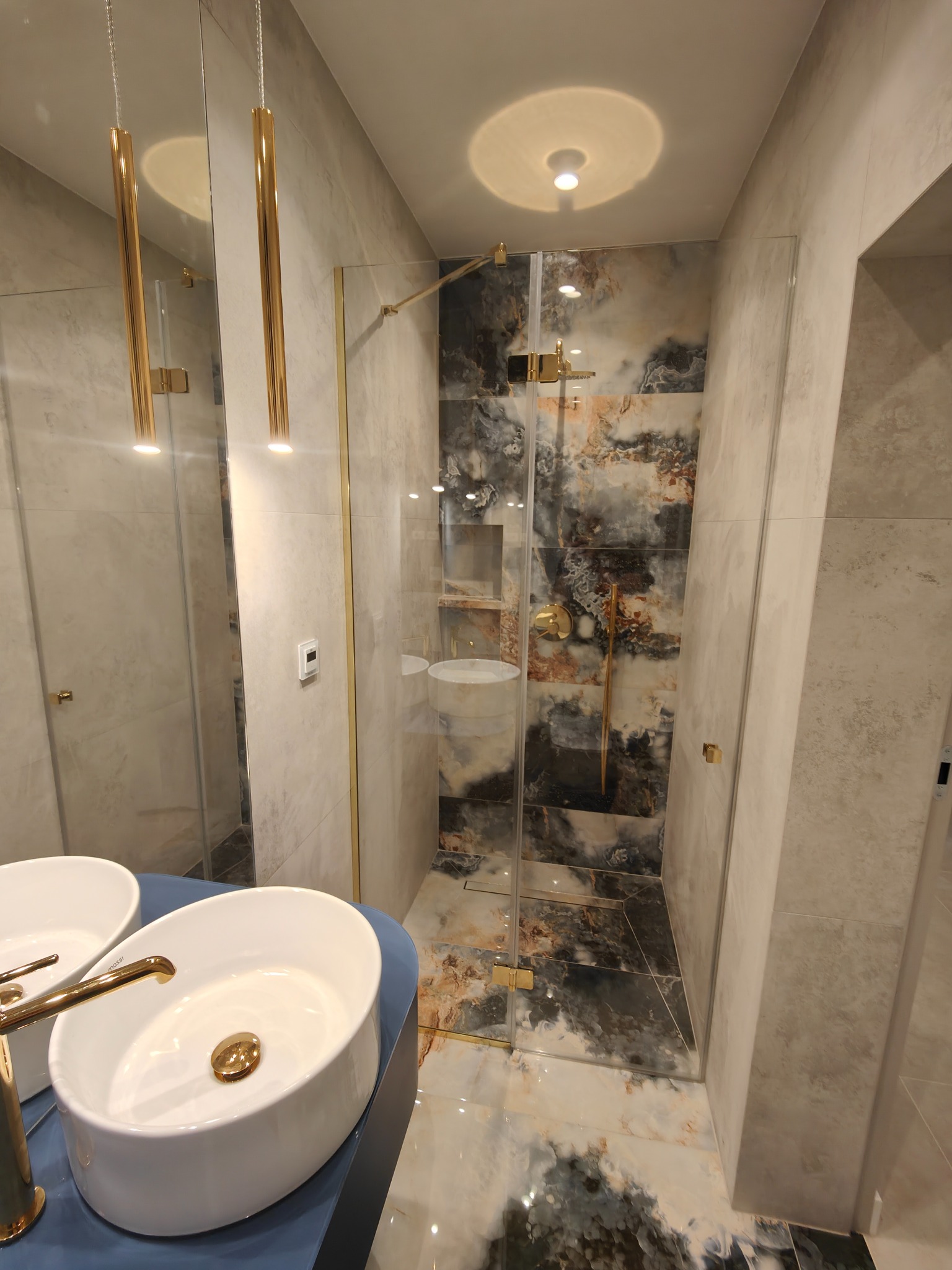 Nowoczesna łazienka z kabiną prysznicową typu walk-in, umywalką nablatową z baterią w kolorze złotym, oraz płytkami imitującymi marmur w odcieniach szarości, bieli i brązu. Na ścianie widoczny...