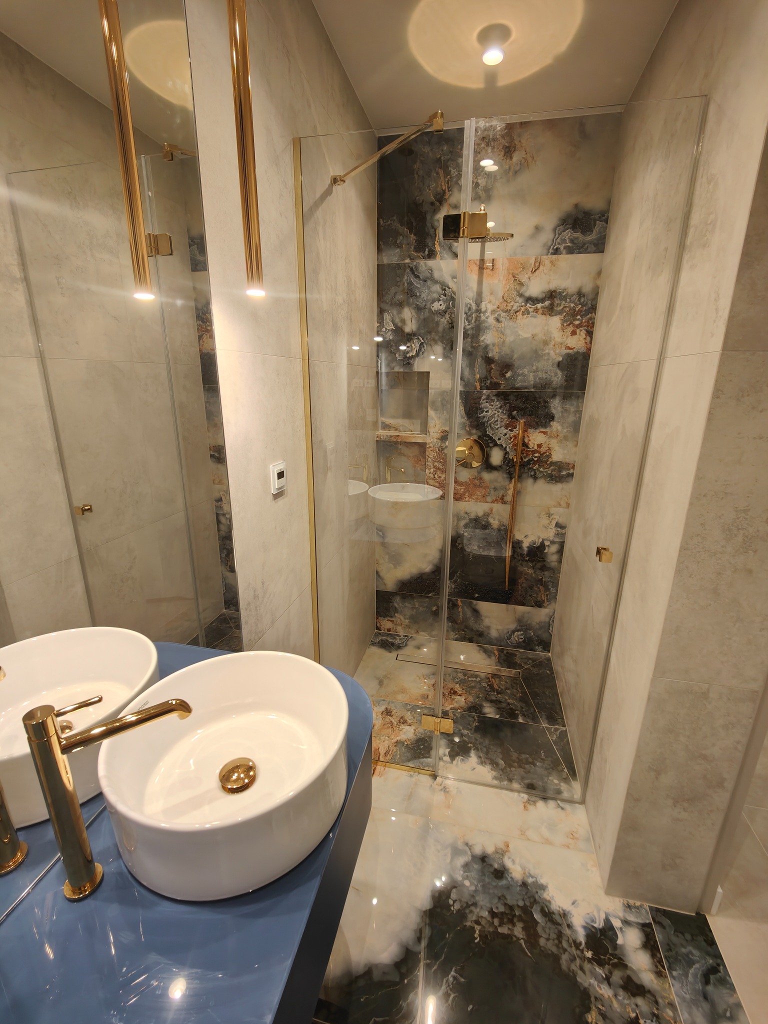 Nowoczesna łazienka z dwoma białymi umywalkami nablatowymi z bateriami w kolorze złotym, kabiną prysznicową ze szklanymi drzwiami i płytkami imitującymi marmur w odcieniach szarości, czerni i złota.