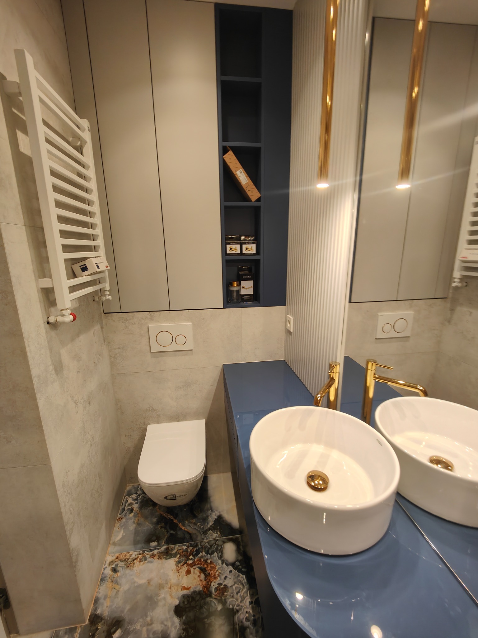 Nowoczesna łazienka z dwiema białymi umywalkami nablatowymi na niebieskim blacie, złotymi bateriami, grzejnikiem drabinkowym i podłogą z marmuru, oświetlona złotymi lampami wiszącymi.