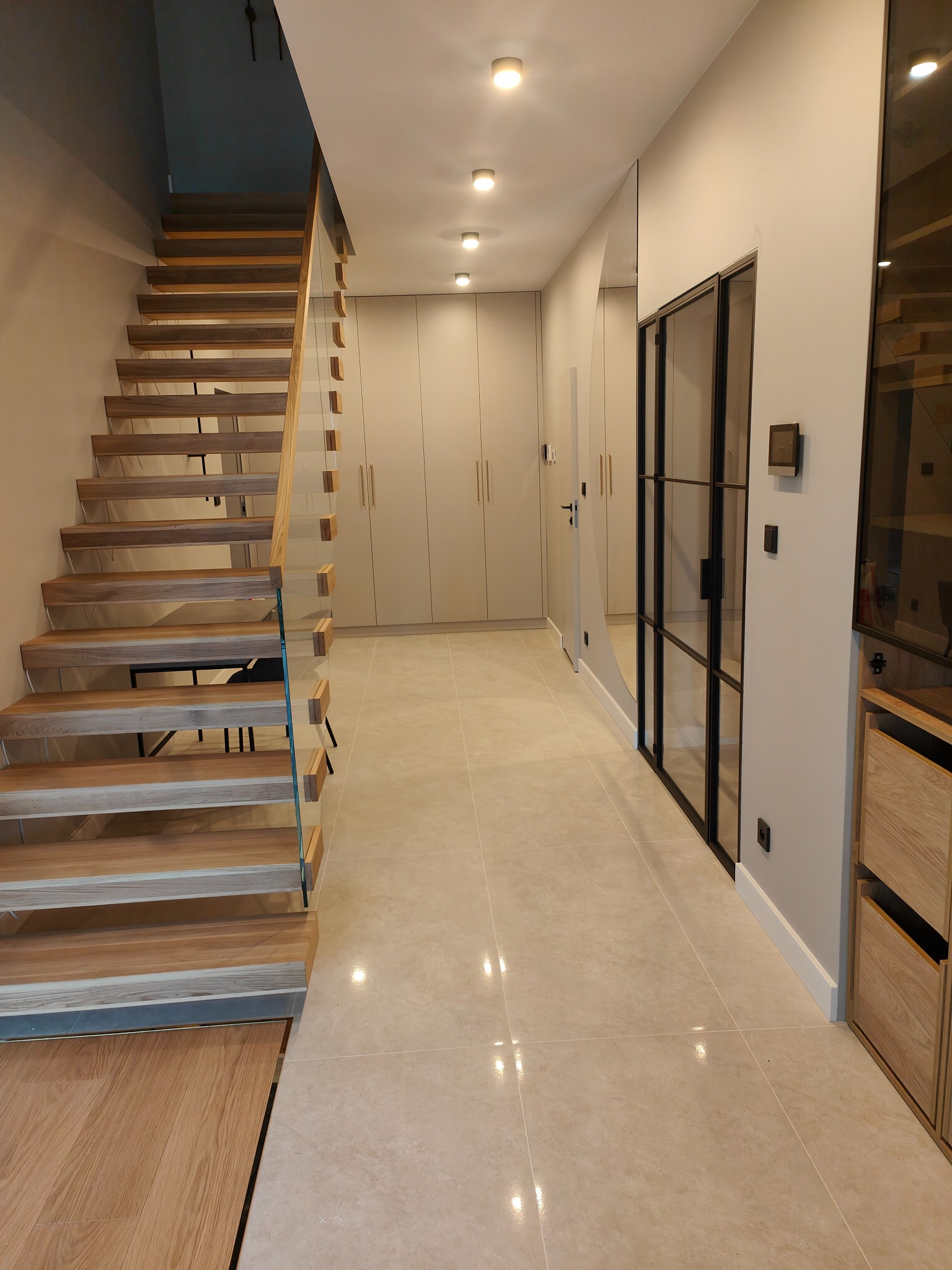 Nowoczesne wnętrze w Żyrardowie z drewnianymi schodami z przezroczystą balustradą, beżową podłogą z płytek, zabudową szaf w kolorze ścian i czarną, przeszkloną ścianką działową.
