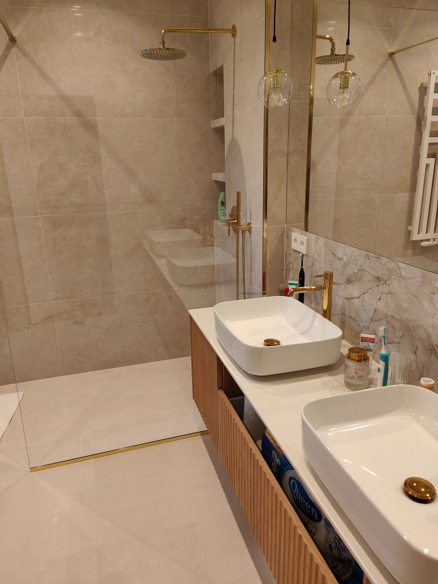 Elegancka łazienka z dwoma białymi umywalkami nablatowymi, złotą armaturą i kabiną prysznicową typu walk-in z subtelnymi złotymi akcentami, oświetlona wiszącymi lampami z żarówkami Edisona.