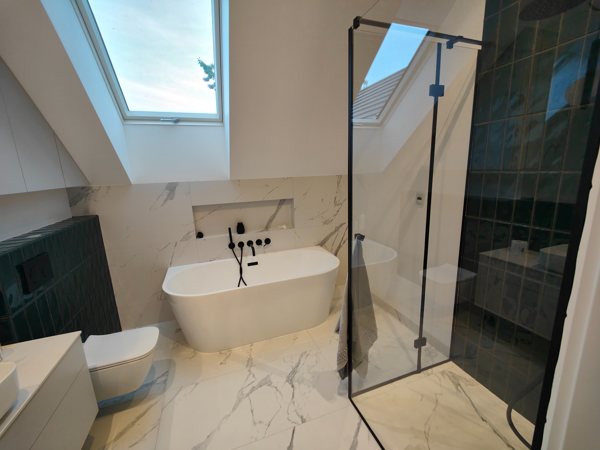 Nowoczesna łazienka na poddaszu z białą wanną wolnostojącą, kabiną prysznicową ze szkła i czarnymi elementami armatury, doświetlona oknem dachowym.