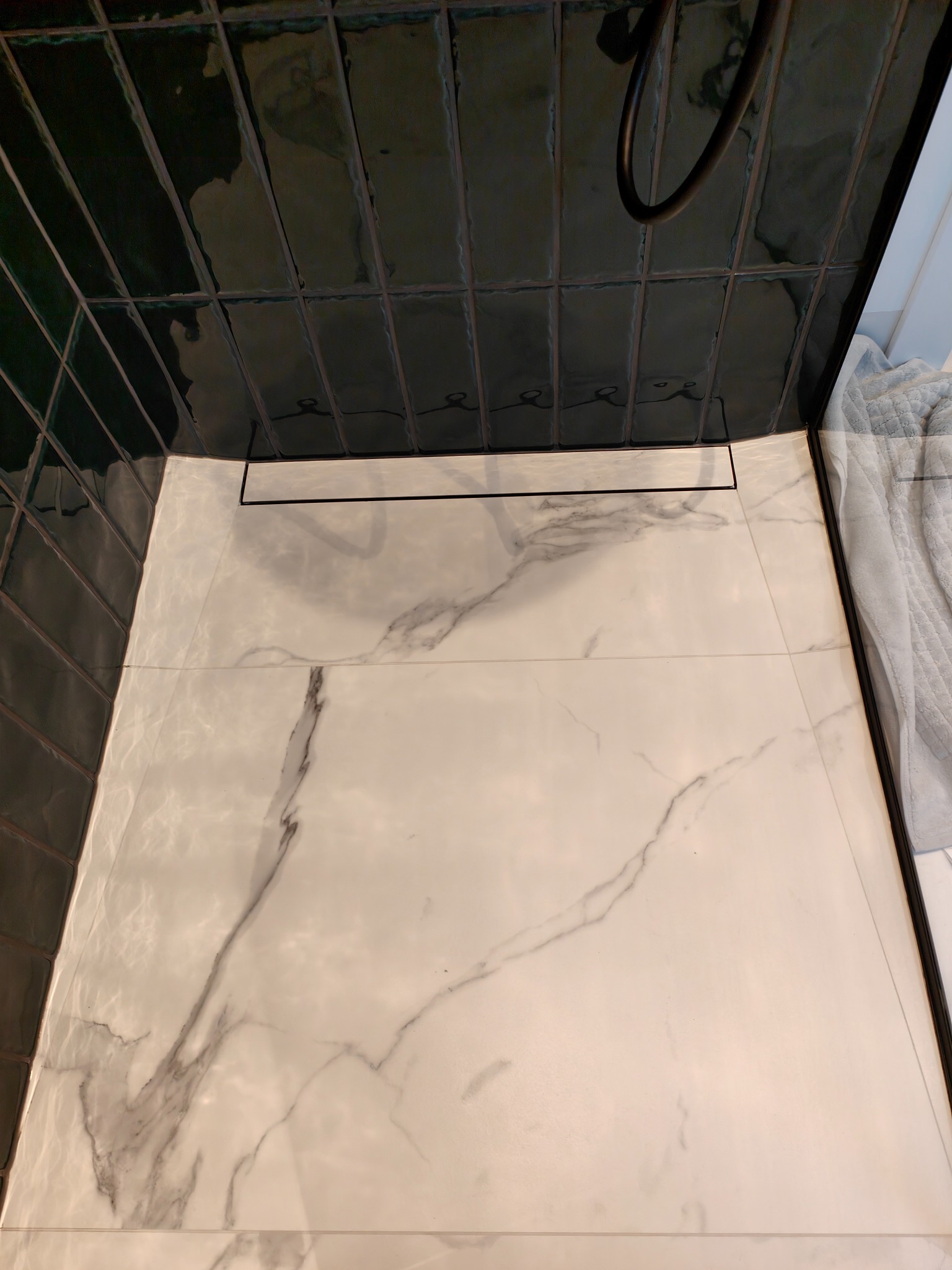 Nowoczesny prysznic walk-in z odpływem liniowym, wykończony białymi płytkami imitującymi marmur i ciemnozielonymi, błyszczącymi płytkami na ścianie.