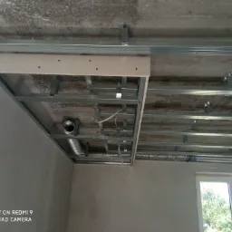 Stelaż sufitu podwieszanego z płyt kartonowo-gipsowych, widoczne profile metalowe, przewody wentylacyjne i fragment płyty z otworami, surowy betonowy strop w tle.