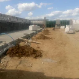 Budowa domu murowanego: widok na plac budowy z częściowo wzniesionymi ścianami z bloczków, przygotowanym fundamentem i stertami ziemi, w tle zieleń drzew.