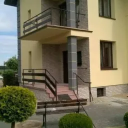 Dwupiętrowy dom z żółtą elewacją, szarymi kolumnami i balkonem, widoczny z poziomu ogrodu z niskimi krzewami i ławką.