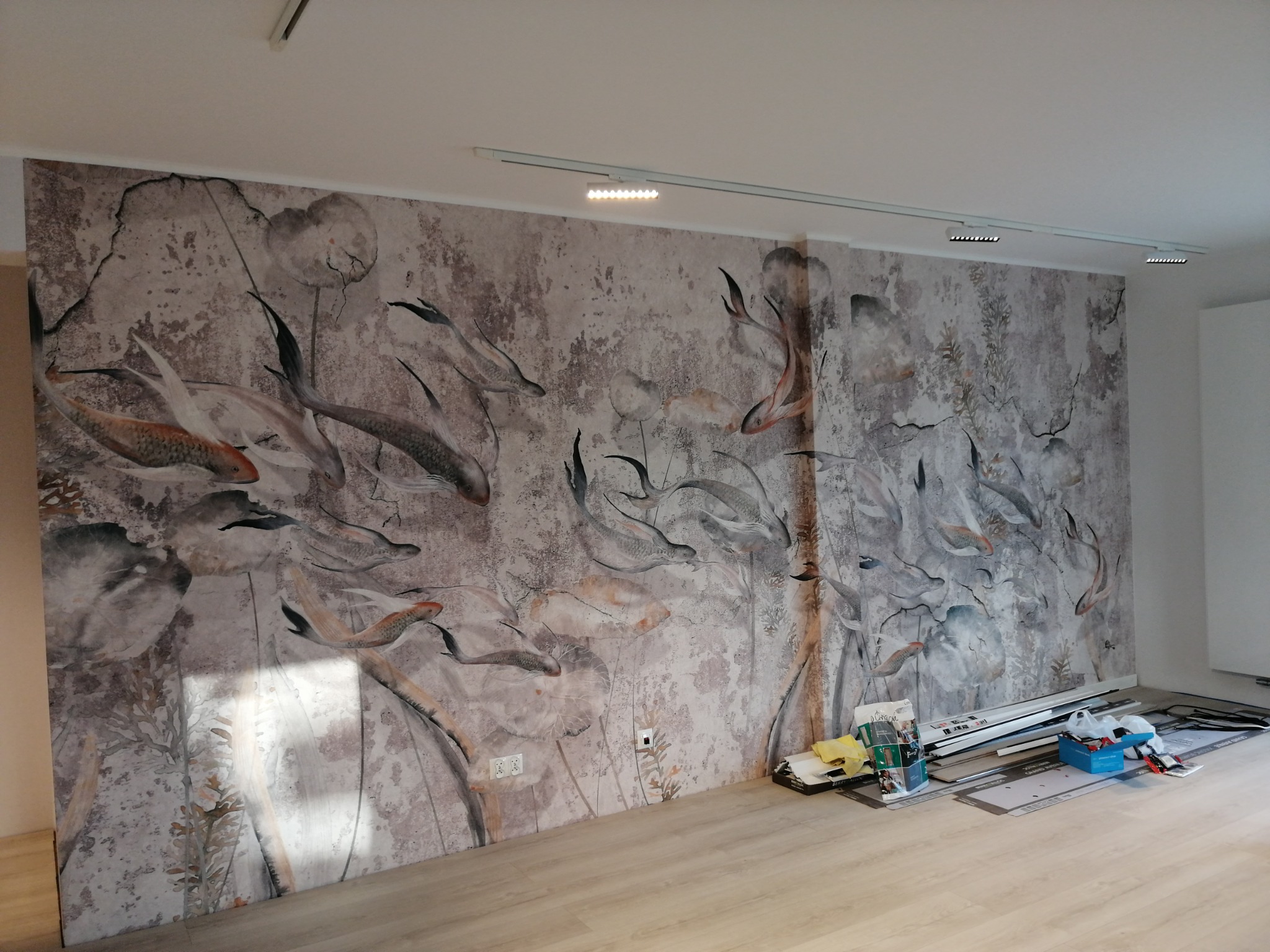 Ściana pokryta fototapetą z motywem ryb Koi i lilii wodnych, widoczne akcesoria do montażu paneli podłogowych, oświetlenie szynowe na suficie.