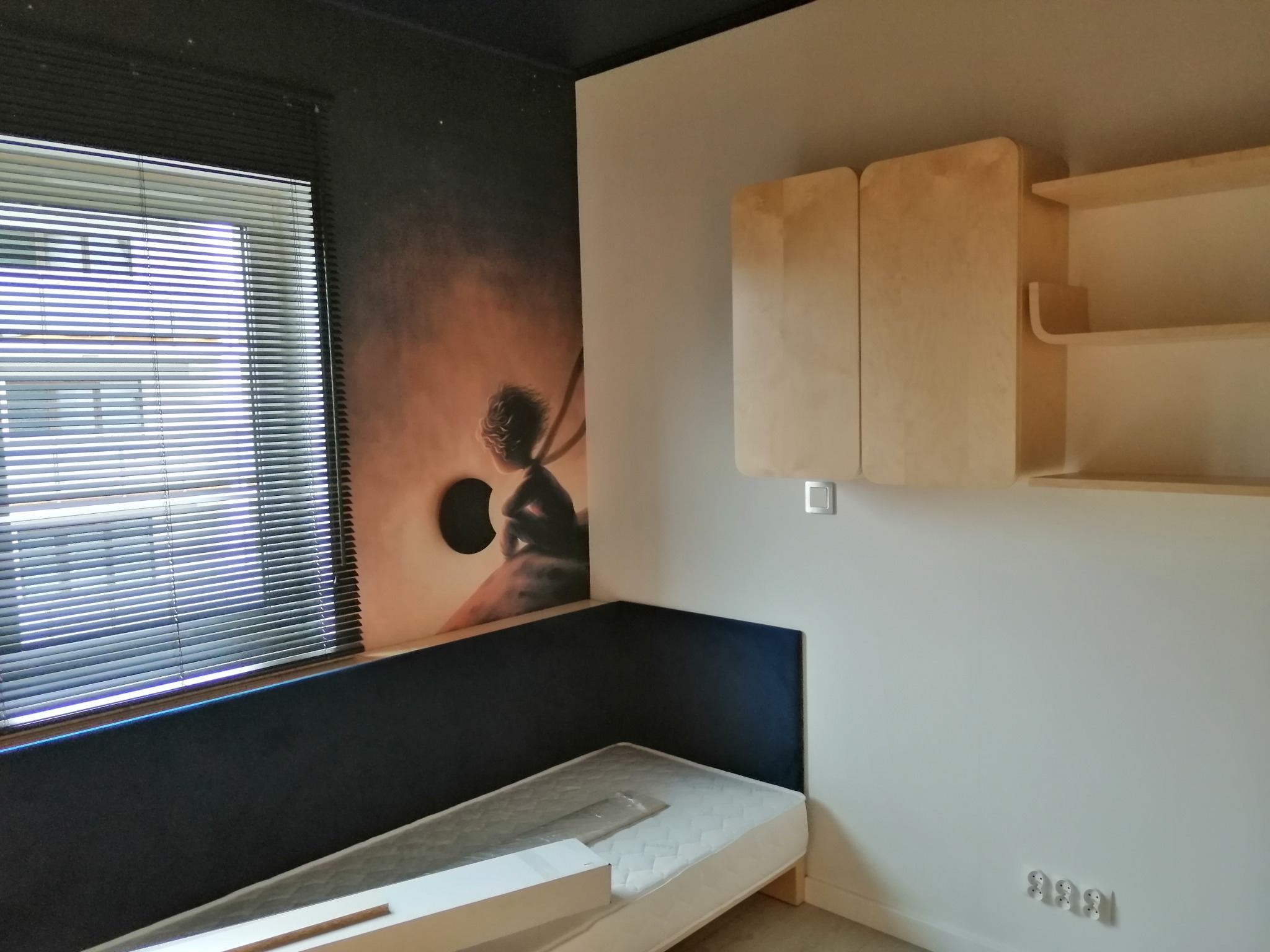 Pokój po remoncie: łóżko z materacem w folii, tapeta z motywem dziecka na huśtawce, zabudowa meblowa z jasnego drewna, rolety okienne.