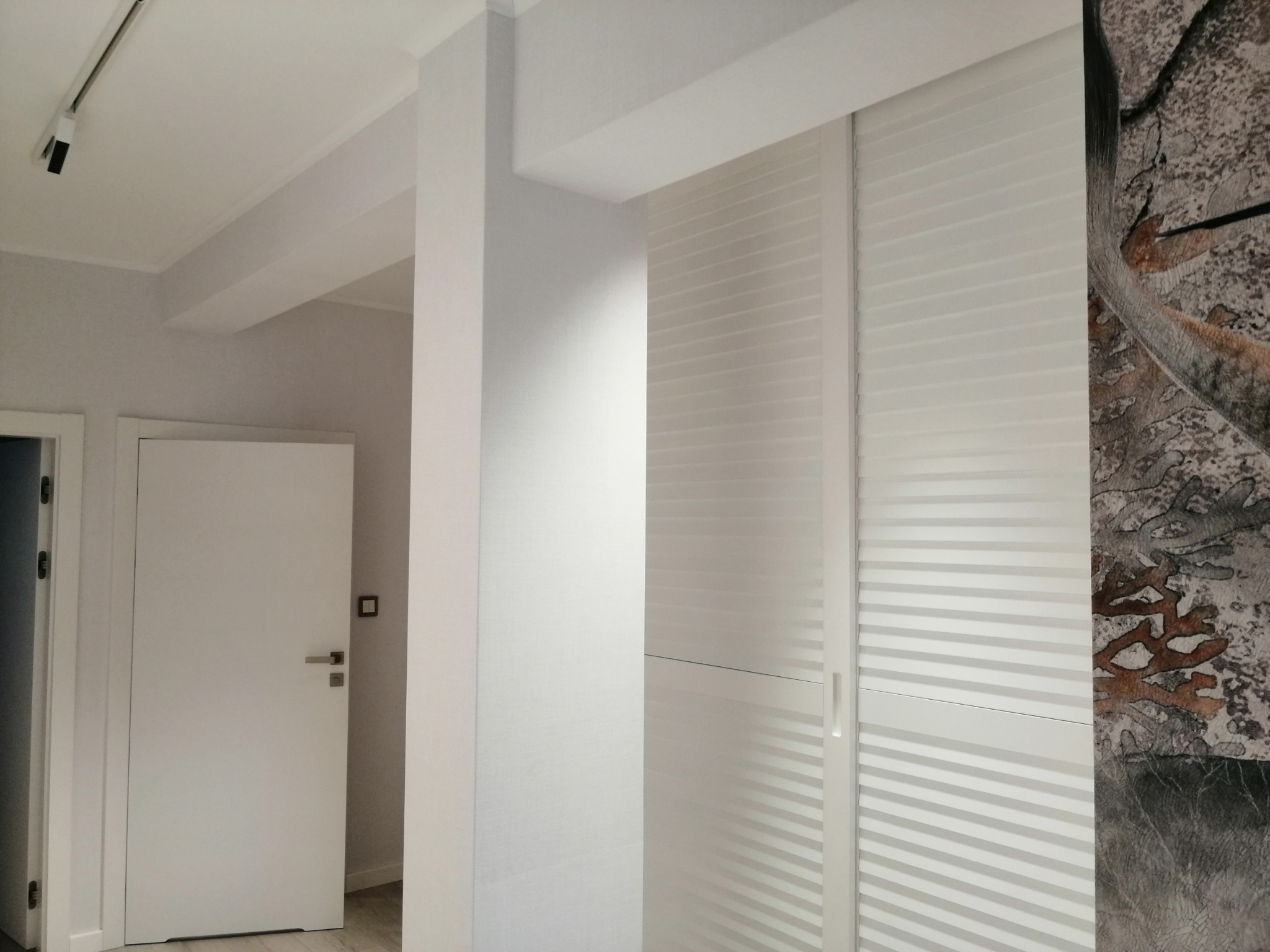 Białe, przesuwne drzwi szafy z poziomymi żaluzjami, przylegające do ściany z abstrakcyjną tapetą w odcieniach szarości i brązu, widoczne również białe drzwi wejściowe i oświetlenie sufitowe.