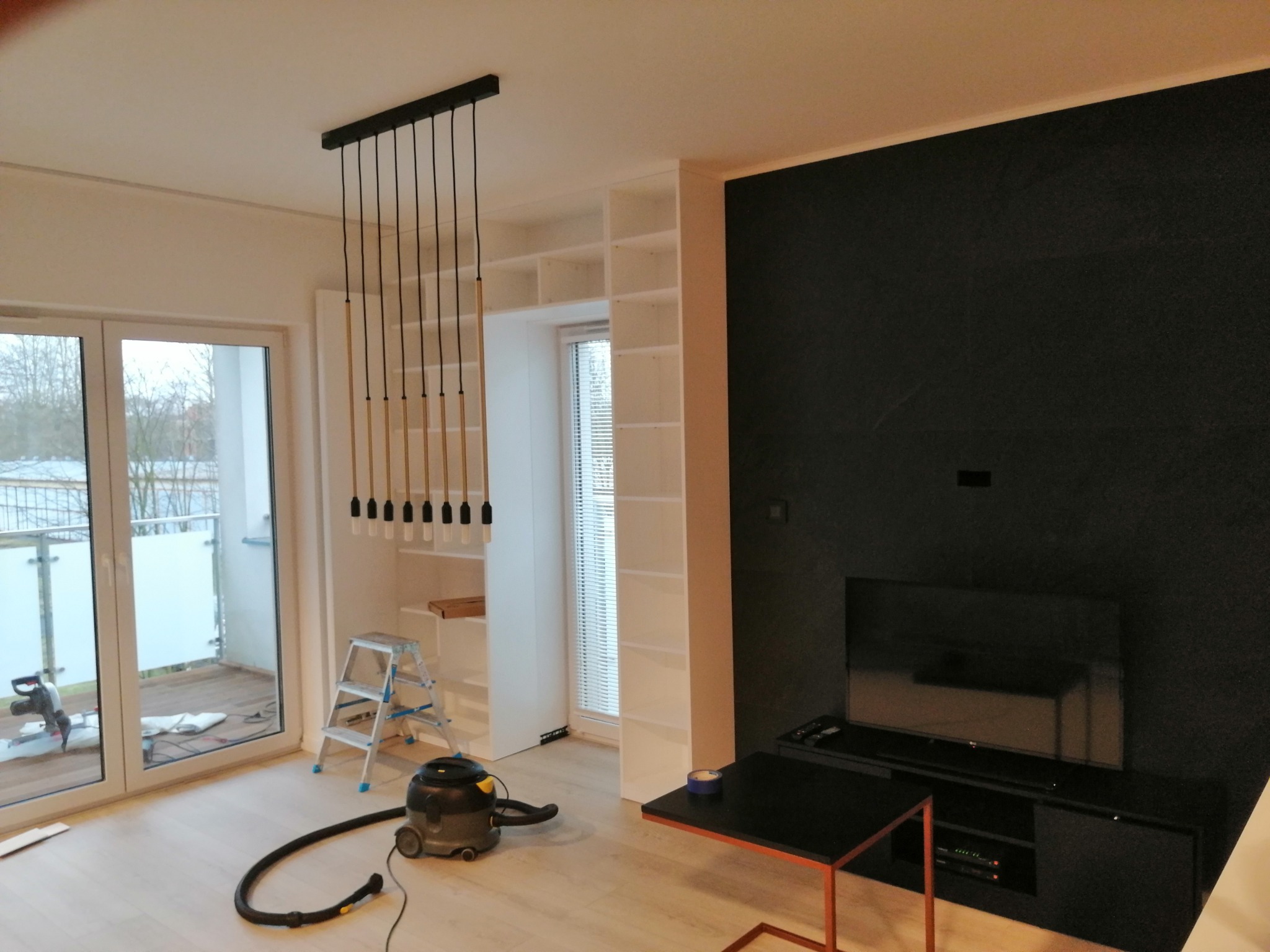 Wnętrze mieszkania w trakcie prac: białe regały w zabudowie, czarna ściana z telewizorem, wisząca lampa z kilkoma żarówkami, drabina i odkurzacz na podłodze.