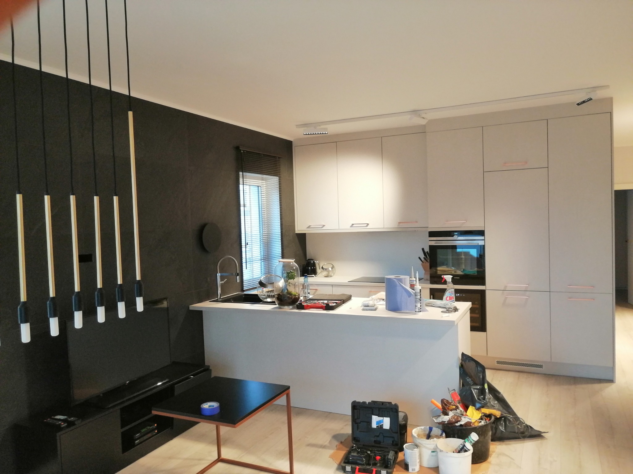 Nowoczesna kuchnia z wyspą w trakcie prac wykończeniowych, widoczne narzędzia i materiały budowlane, minimalistyczne lampy wiszące na tle ciemnej ściany z telewizorem.