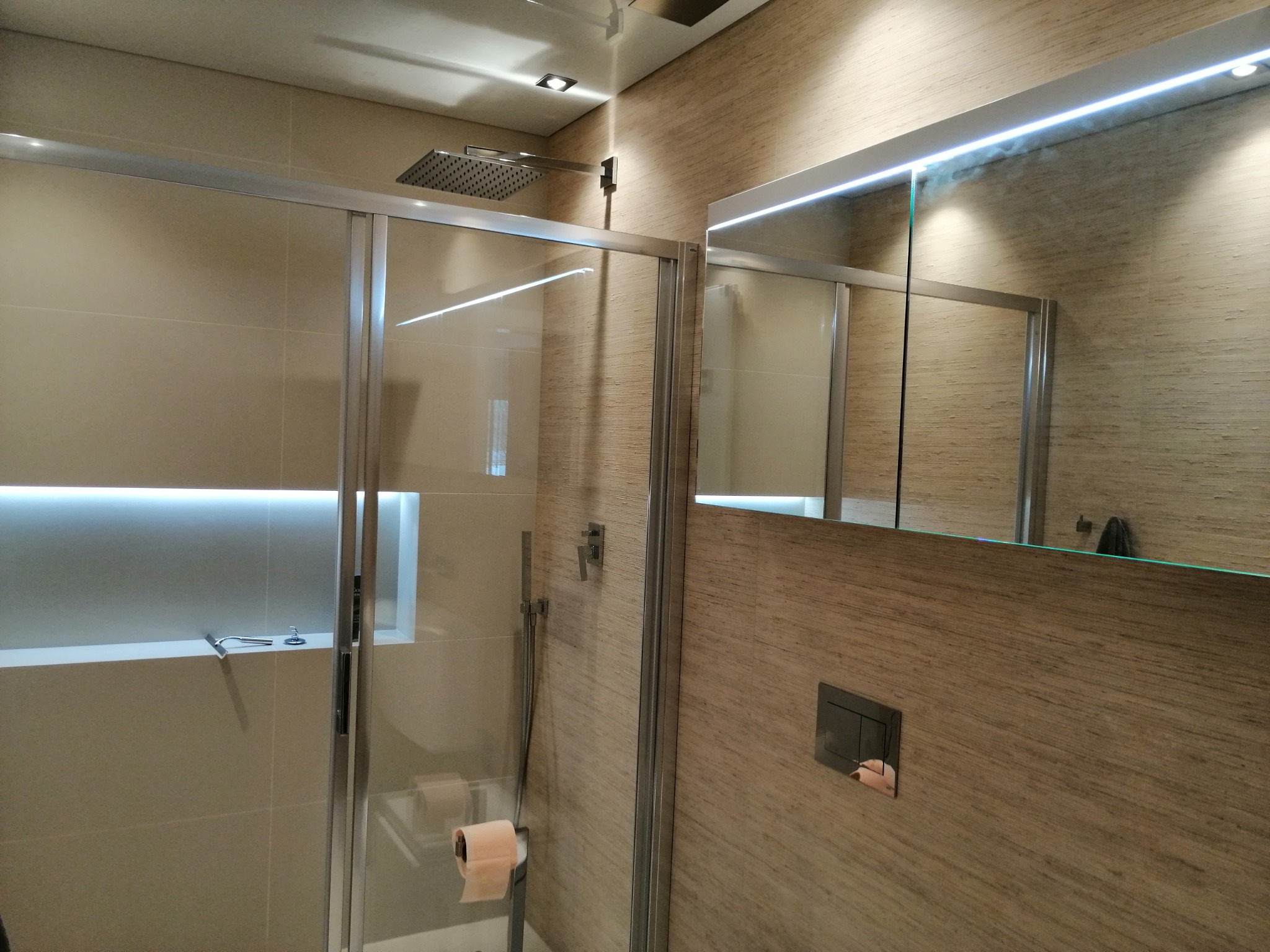 Nowoczesna łazienka z kabiną prysznicową typu walk-in, deszczownicą i podświetlanym lustrem, wykończona płytkami imitującymi drewno i z ukrytym oświetleniem LED.