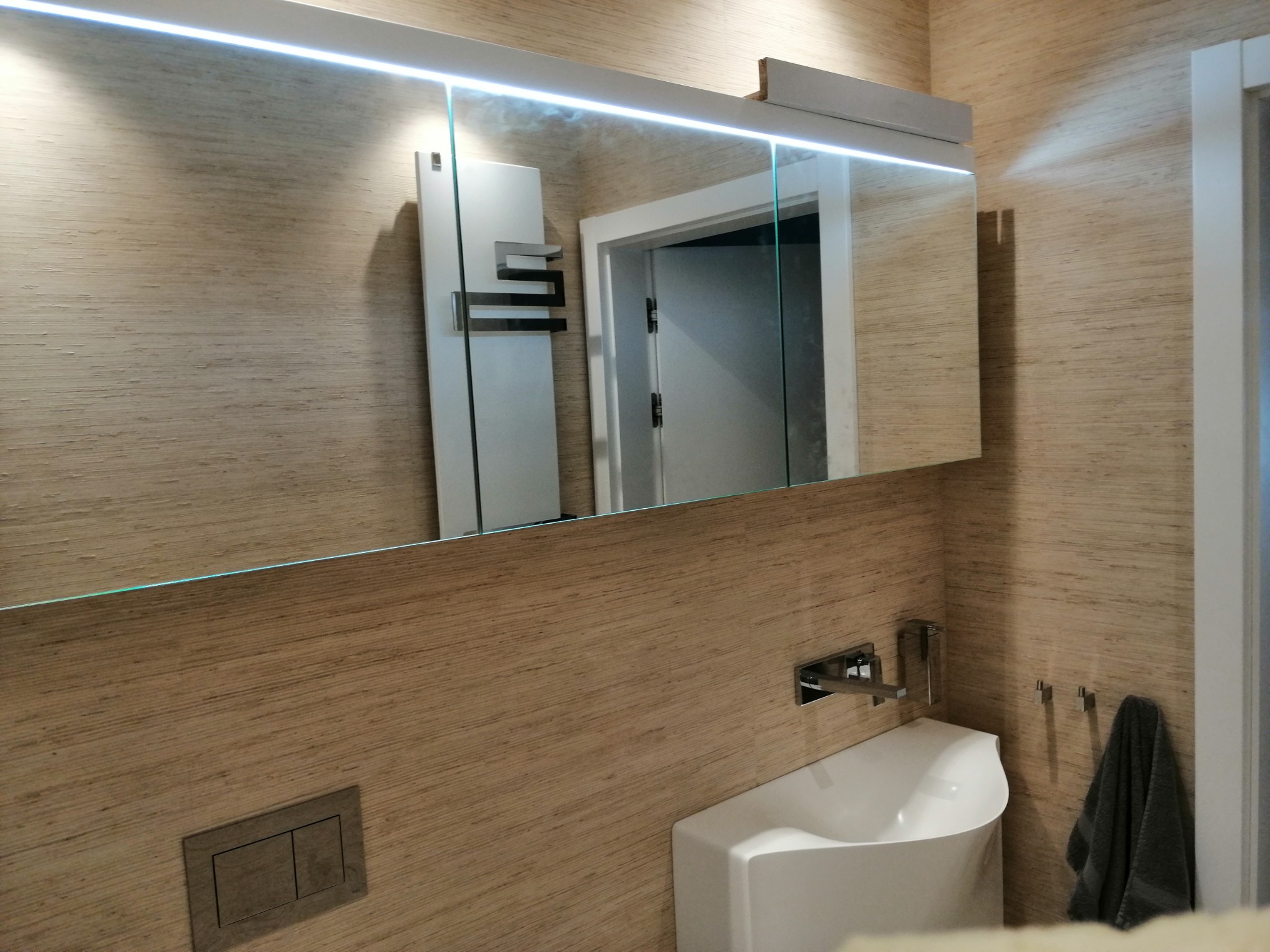 Nowoczesna łazienka z umywalką narożną, lustrem z oświetleniem LED i tapetą o fakturze drewna.