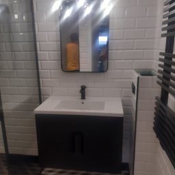 BAS Usługi Remontowo-Budowlane - Nowoczesna łazienka z białą cegiełkową glazurą, czarną szafką pod umywalką, lustrem z zaokrąglonymi rogami i trzema wiszącymi lampami, oraz grzejnikiem drabinkowym w kolorze czarnym.