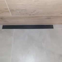 Ułożona podłoga z szarych płytek w łazience, z odpływem liniowym w kolorze czarnym, przy ścianie wykończonej płytkami imitującymi drewno.