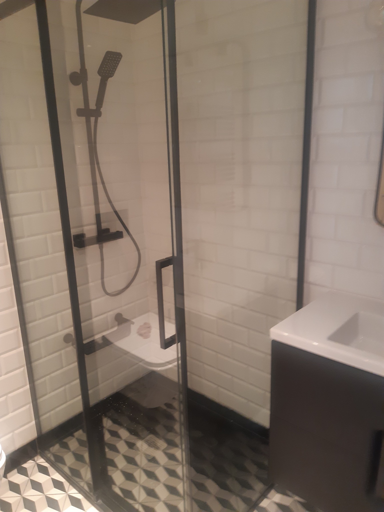 Nowoczesna kabina prysznicowa z czarną ramą i deszczownicą, białe płytki imitujące cegłę na ścianach, wzorzysta podłoga, umywalka z szafką w tle.