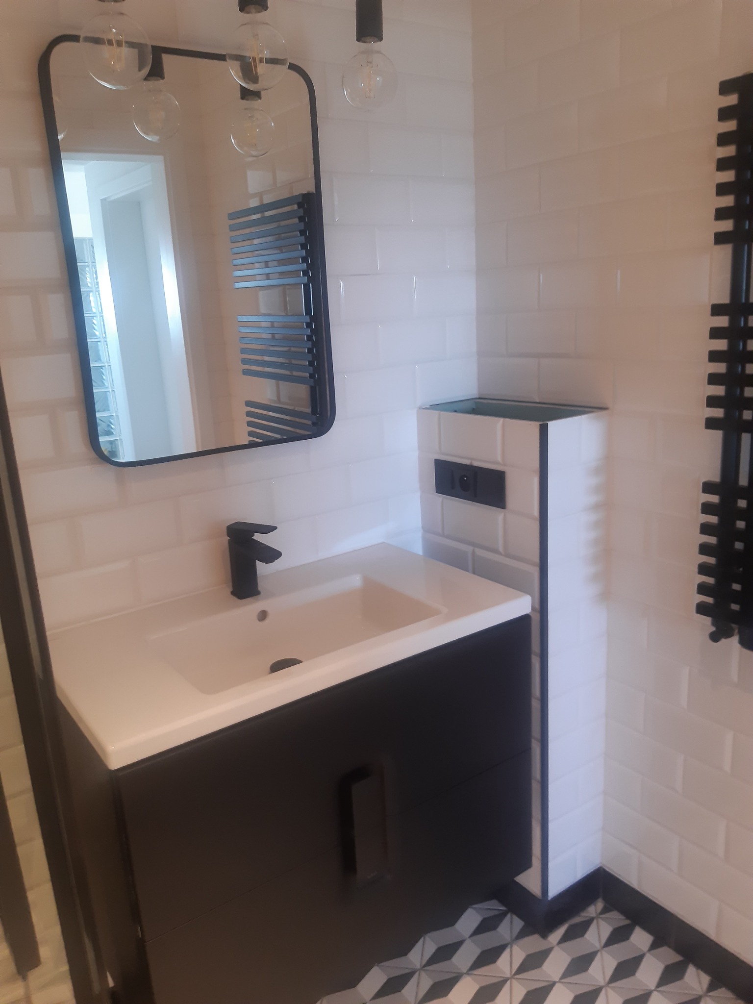 Nowoczesna łazienka z białą armaturą, czarną szafką pod umywalką i geometryczną podłogą. W tle lustro z czarną ramą i grzejnik drabinkowy.