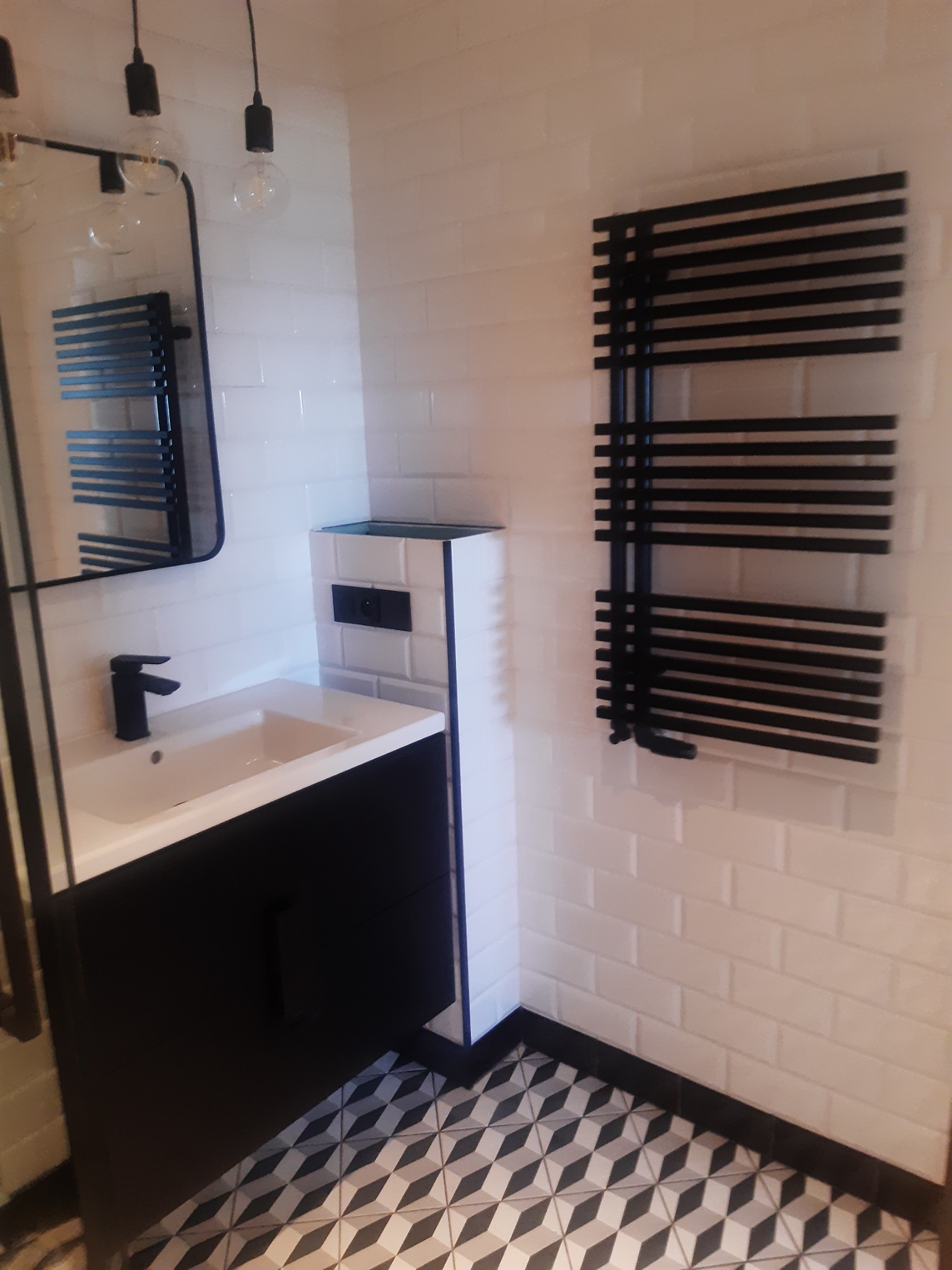Nowoczesna łazienka z białą cegiełkową okładziną, czarnym grzejnikiem drabinkowym, szafką umywalkową w kolorze czarnym i geometryczną podłogą w odcieniach szarości i czerni.
