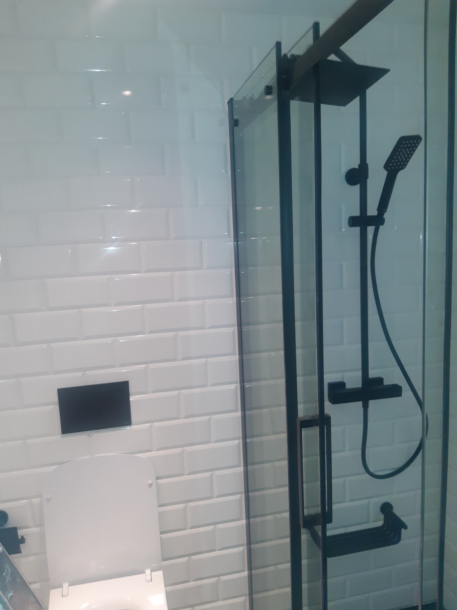 Nowoczesna łazienka z białą, kafelkową ścianą typu metro, czarną armaturą prysznicową za szklaną kabiną oraz toaletą z czarnym przyciskiem spłuczki.