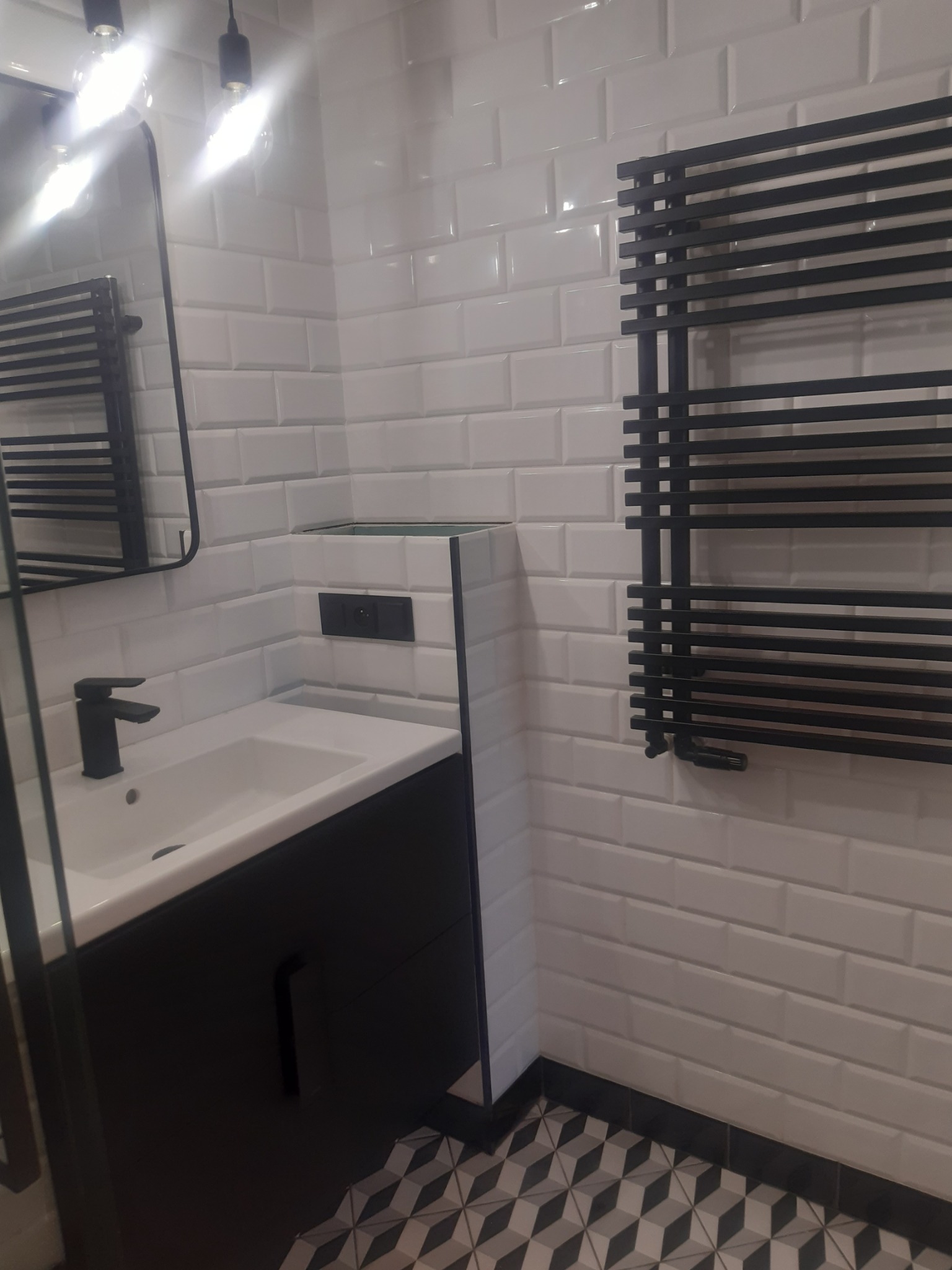 Nowoczesna łazienka z białą cegiełkową glazurą, czarną armaturą, grzejnikiem drabinkowym i geometryczną podłogą.