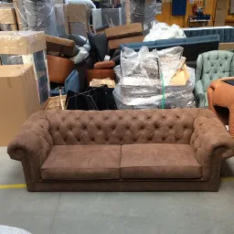 Brązowa sofa Chesterfield z guzikami, otoczona meblami w magazynie lub warsztacie, w tle kartony i folie ochronne.