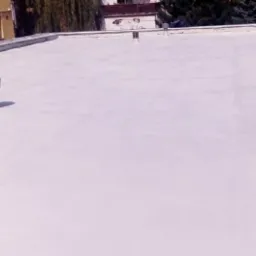 Biała, gładka powierzchnia dachu po hydroizolacji, widoczne kominki wentylacyjne i budynek w tle.