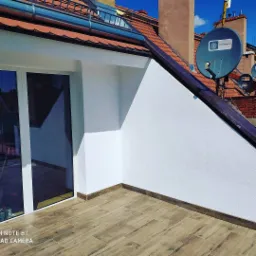 Biała elewacja na balkonie z drewnopodobną podłogą, widok na dach z antenami satelitarnymi, błękitne niebo w tle.