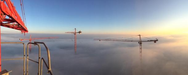 Panorama z wysokości dźwigu: trzy żurawie budowlane wystające z morza mgieł oświetlonych wschodzącym słońcem.
